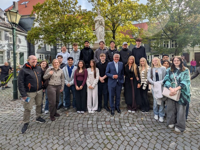 Gruppenfoto der Klasse HW 263 mit Bundespräsident Frank-Walter Steinmeier in der Mitte, dazu Lehrkraft und Schulleitung, vor einem Brunnen mit Frauenstatue zwischen Fachwerk- und Schieferhäusern in der Hattinger Altstadt