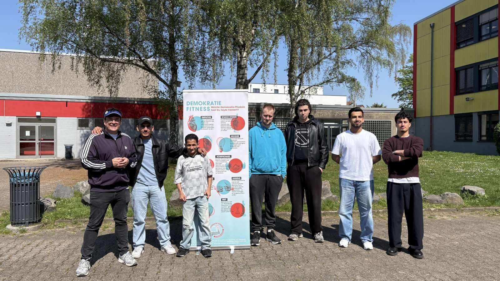Sieben Lernende stehen neben einem Rollbanner mit der Aufschrift Demokratie und Fitness auf dem Schulhof