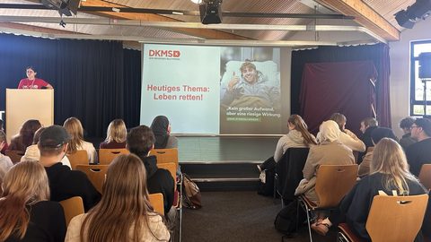 Vortrag im Forum: Eine Ehrenamtliche der DKMS steht am Rednerpult, auf der Leinwand die Folie „Heutiges Thema: Leben retten!“, im Vordergrund Schüler:innen auf Stuhlreihen