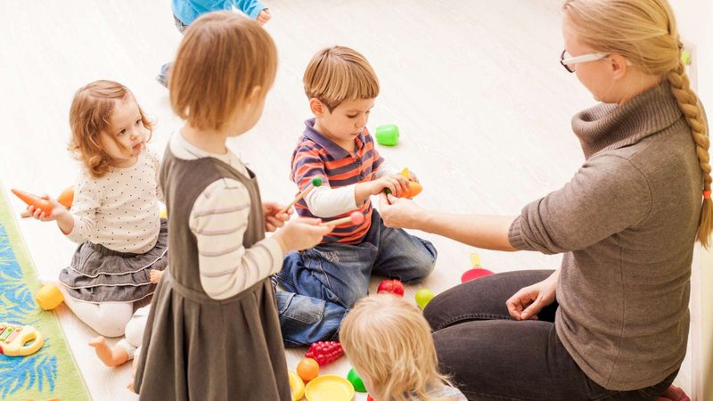 Eine Erzieherin sitzt mit vier Kindern auf dem Boden und spielt mit Obstattrappen
